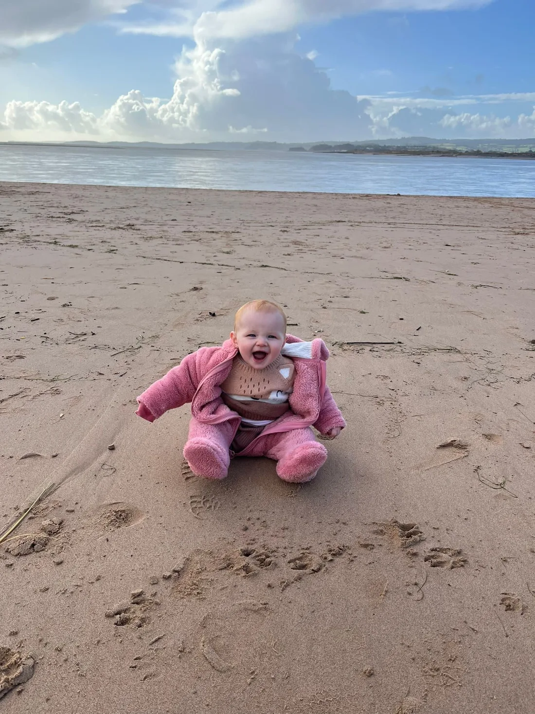 Elsie as a baby, sitting on a beach in a pink suit, laughing